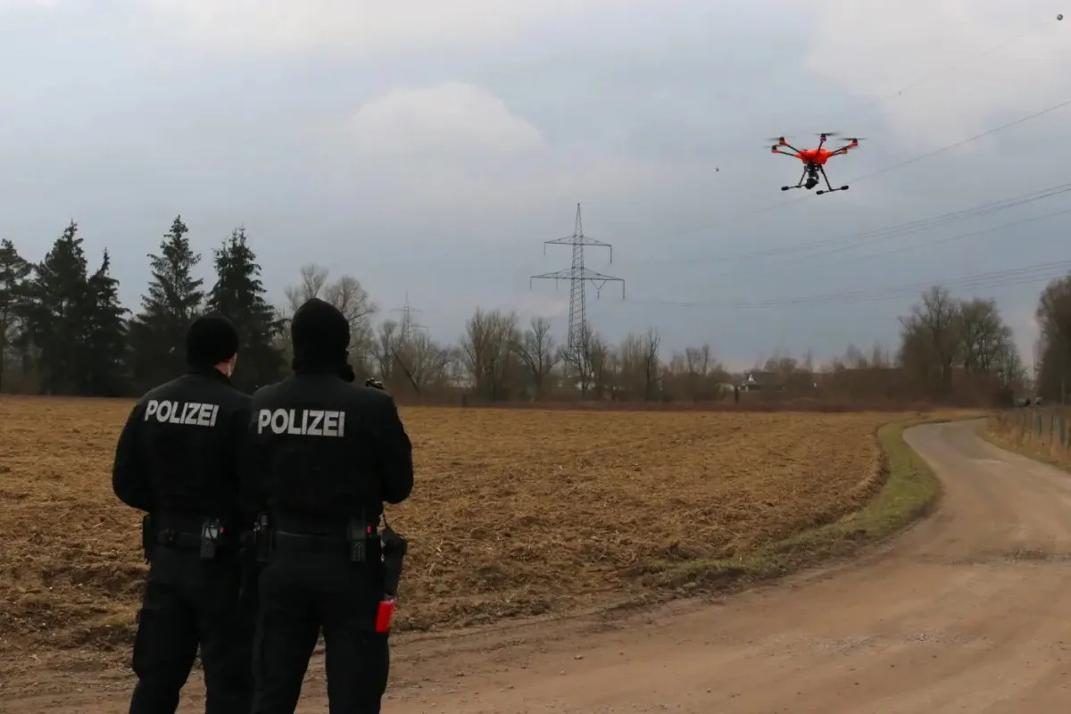 Zwei Polizist*innen stehen vor einem gepflügten Feld, vor ihnen fliegt ein Multicopter/Drohne.